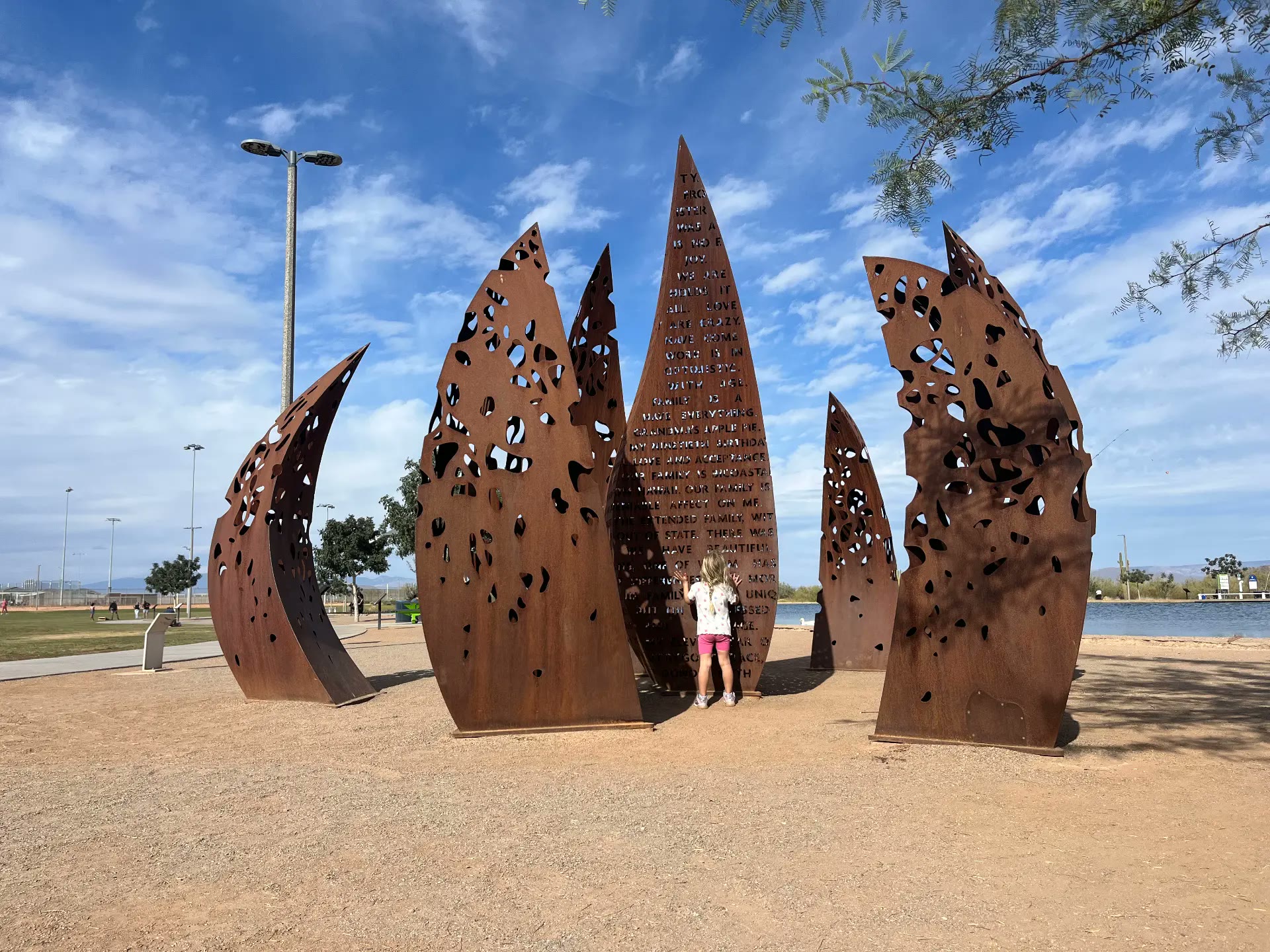 Weathered steel sculpture of pierced leaf forms in a desert park, with a child reaching toward the engraved center panel